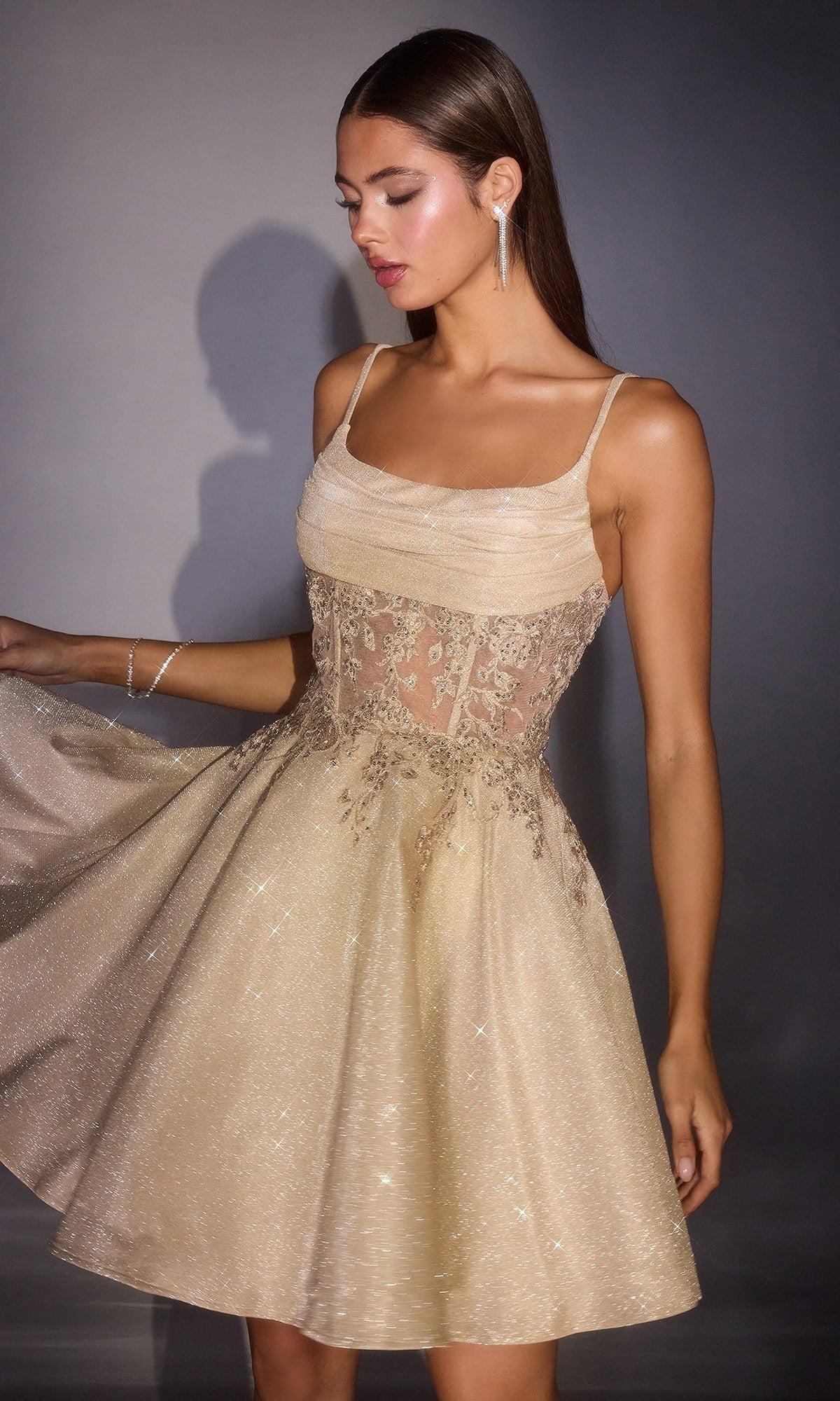 A woman in the Ladivine KV1152 short homecoming dress—with a sleeveless gold fit and flare design, sheer lace bodice, and glitter A-line skirt—poses against a gray background.